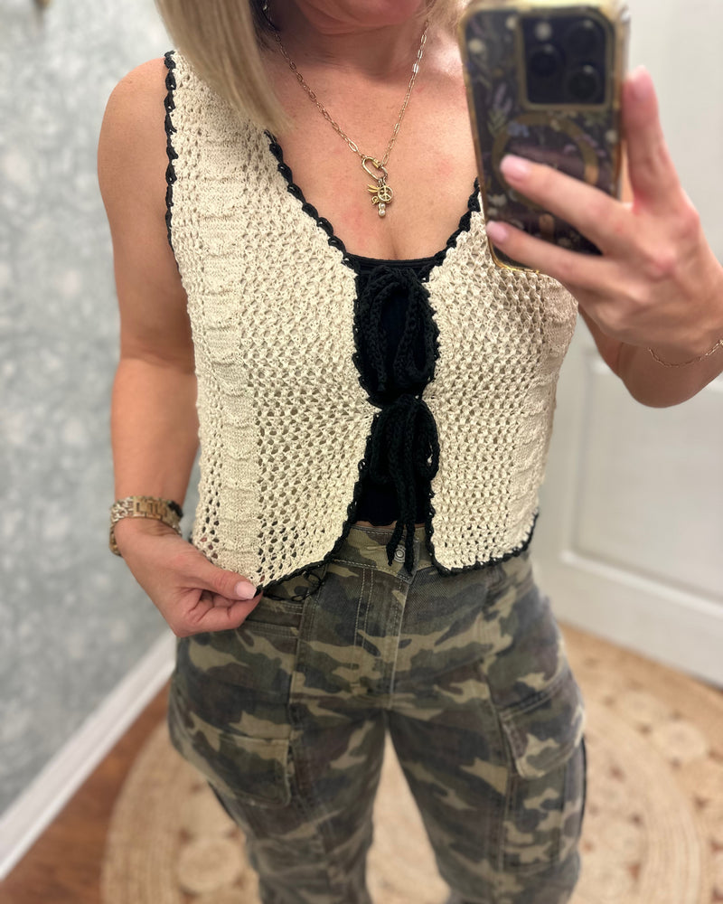Cream crochet vest trimmed with black woven ribbon that front ties into bows. Paired with camo cargo pants.