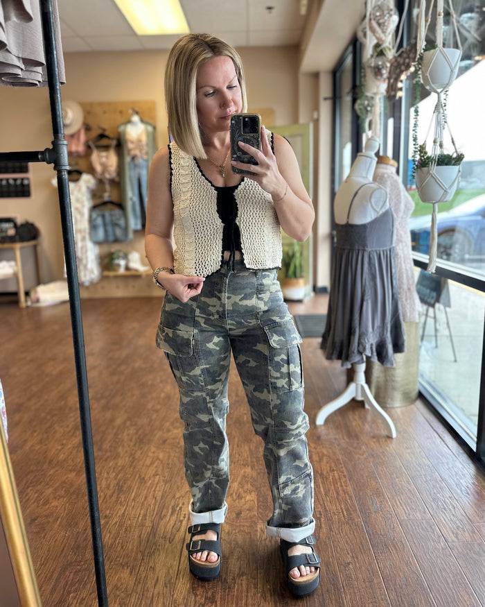 Cream crochet vest trimmed with black woven ribbon that front ties into bows. Paired with camo cargo pants and black sandals.